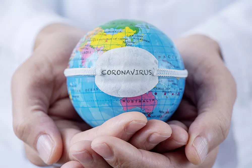 Globe held in cupped hands wearing a small mask labeled “CORONAVIRUS.
