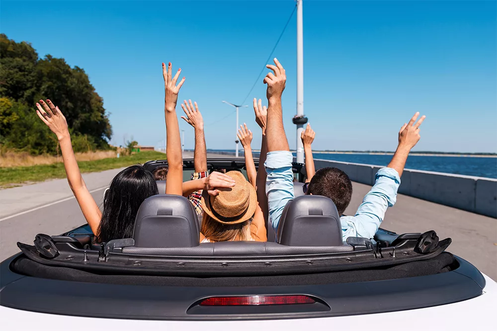 Friends in a white convertible raise hands on coastal road trip under clear blue sky, joyful summer drive.