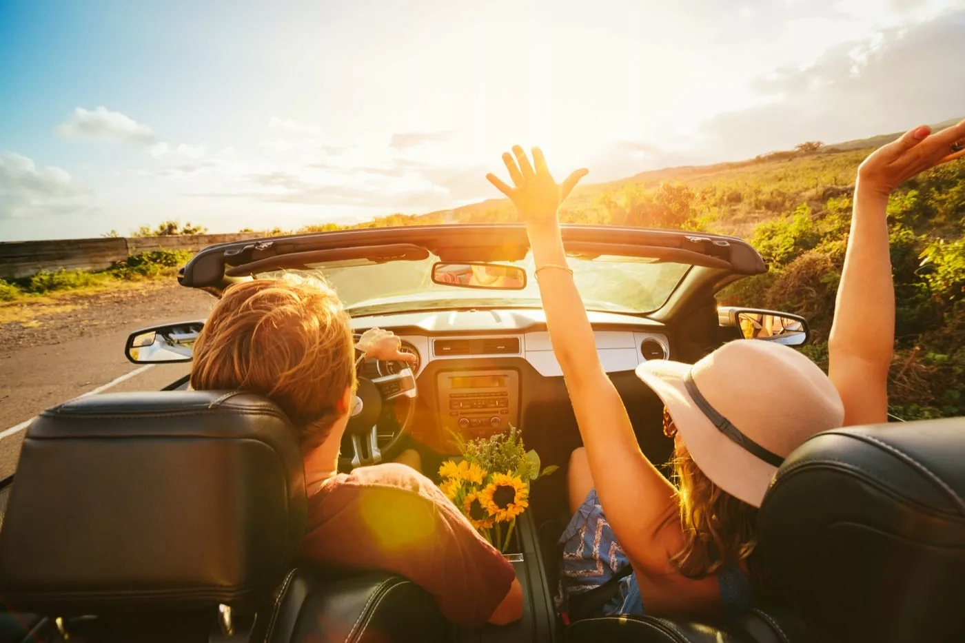 Couple in convertible on sunny coastal road; passenger raises arms, sunflowers between seats. Couple in convertible on sunny coastal road; passenger raises arms, sunflowers between seats.