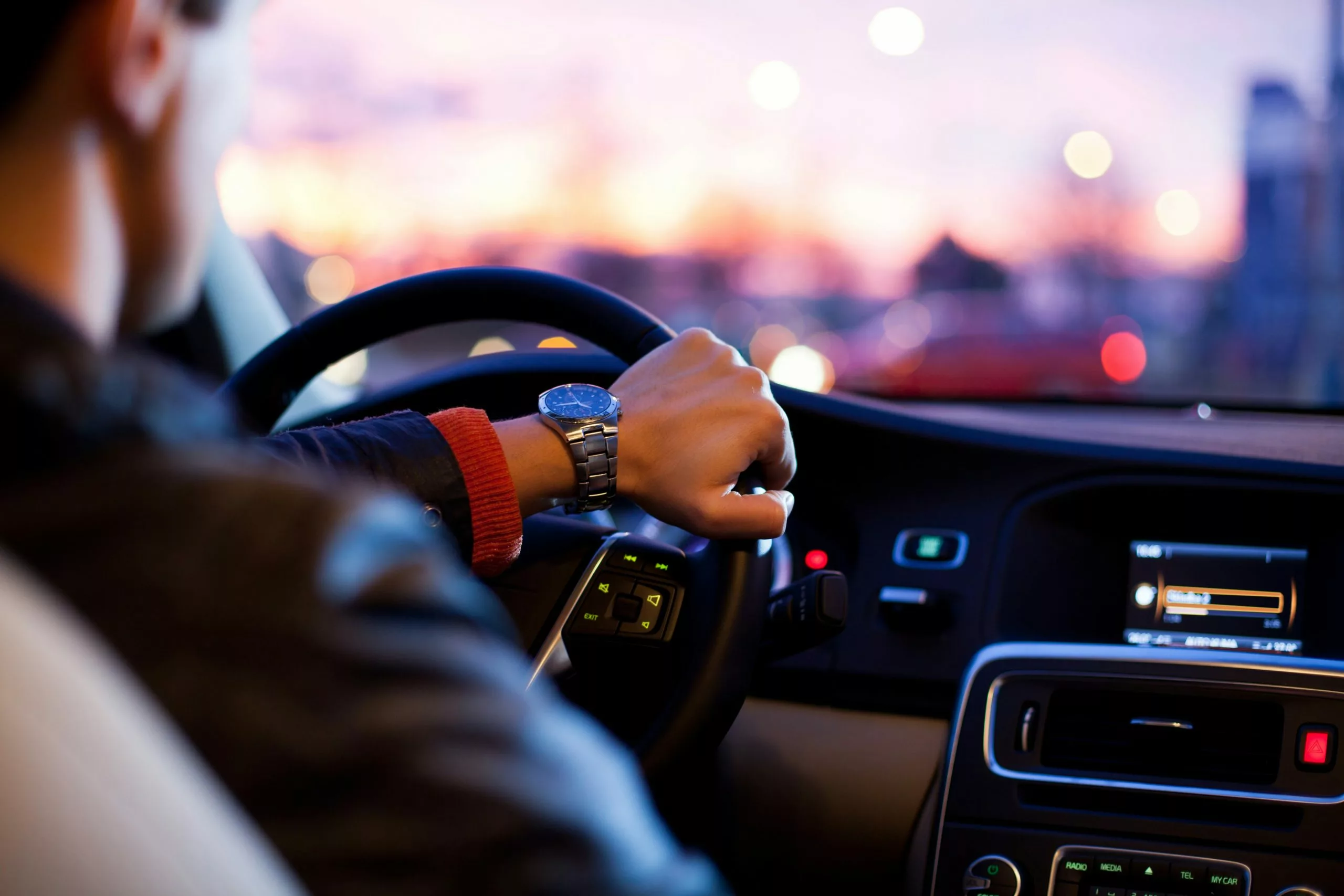 Driver's hand on steering wheel at dusk, city lights blurred outside — Toronto car rental scene.