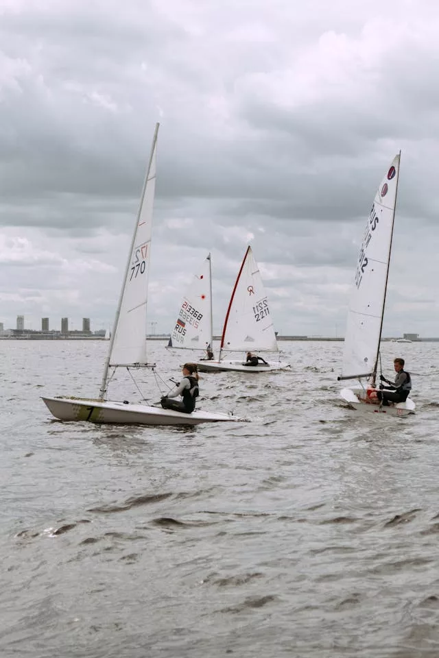 Sailboats racing near waterfront with sailors in small dinghies, cloudy sky and distant city skyline.