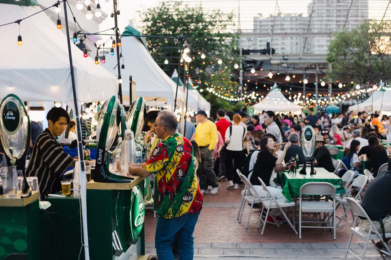 Outdoor food festival with crowded stalls, string lights, people eating and vendors serving drinks.