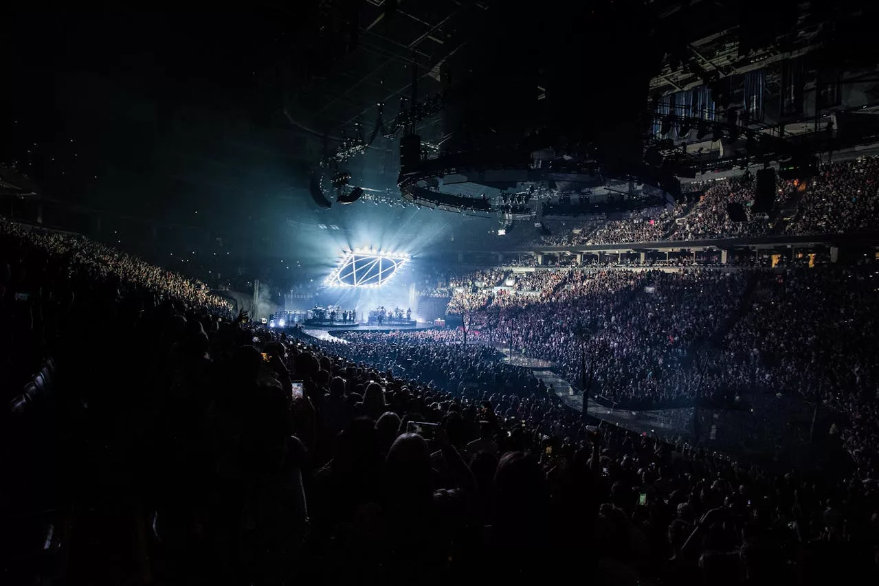 Packed arena concert at night with bright stage lights and a large cheering crowd.